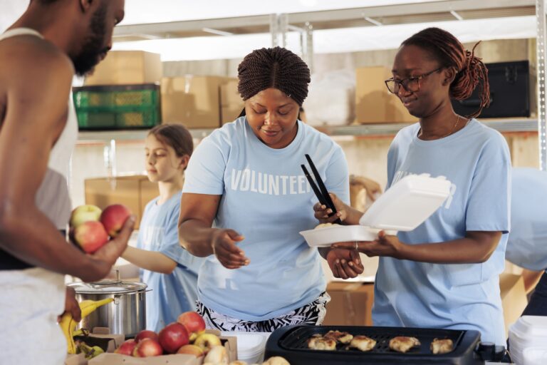 Friendly black women happily serving free food to the poor and needy african american man at a homeless shelter. Hungry, less fortunate male individual receives meal donation from hunger relief team.